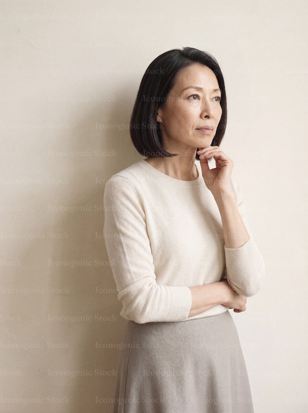 Thoughtful mature Asian woman in cream sweater standing against neutral wall, looking away