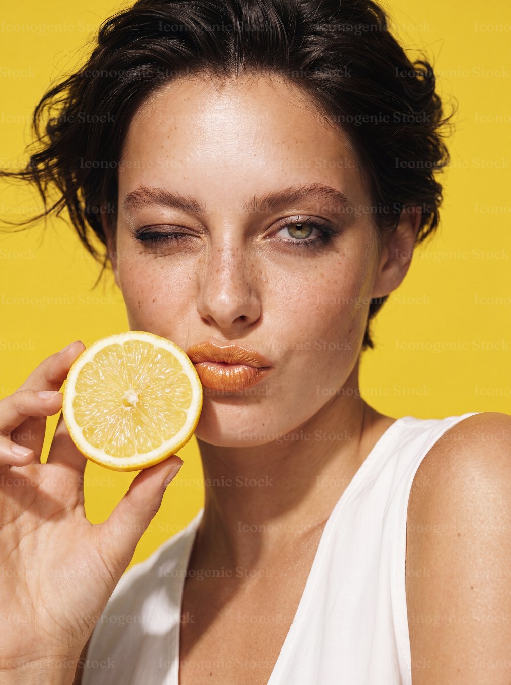 Playful woman winking with pouted lips holding lemon slice on yellow background