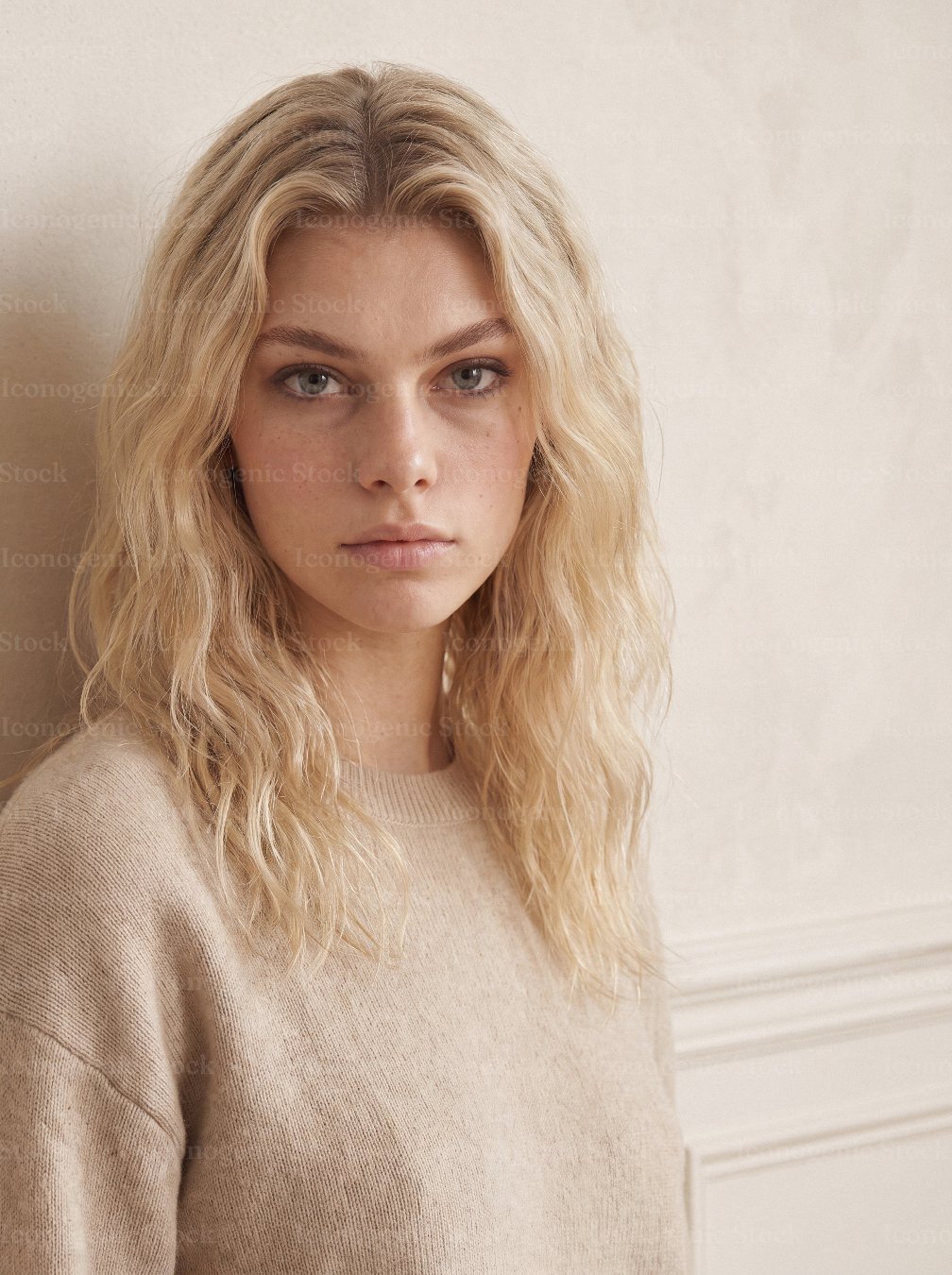 Blonde woman with wavy hair and natural makeup wearing beige sweater, neutral studio portrait