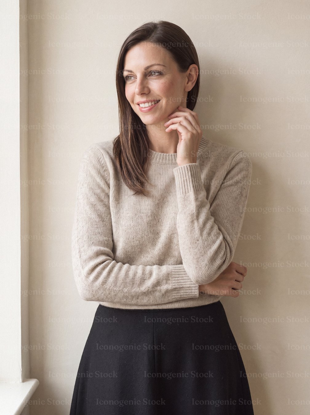 Smiling woman in beige sweater standing by window, looking away thoughtfully
