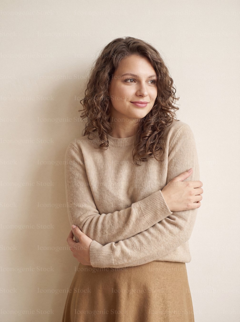 Young woman with curly hair smiling, arms crossed, wearing beige sweater against neutral wall