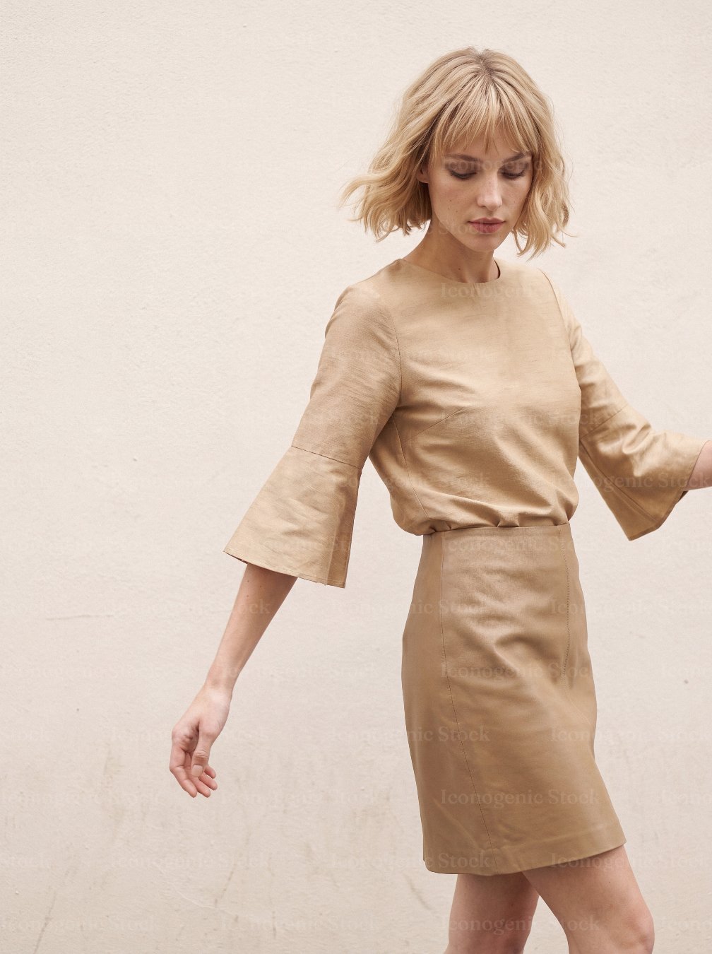 Blonde woman in beige bell-sleeve blouse and leather skirt walking against wall