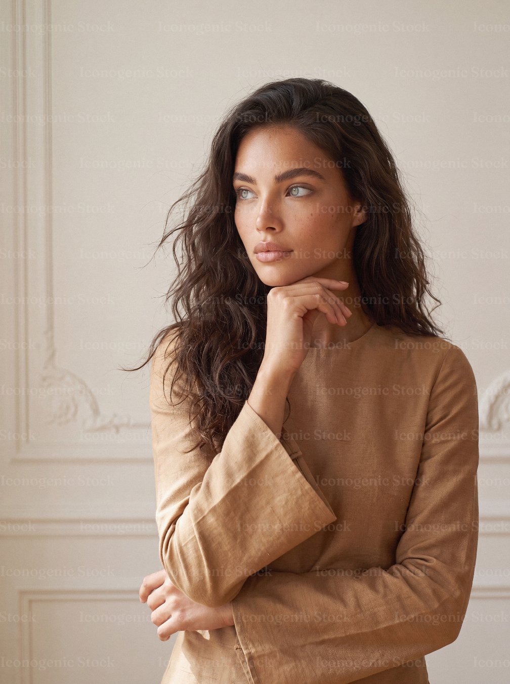 Thoughtful young woman with wavy hair in beige linen blouse, hand on chin, looking away