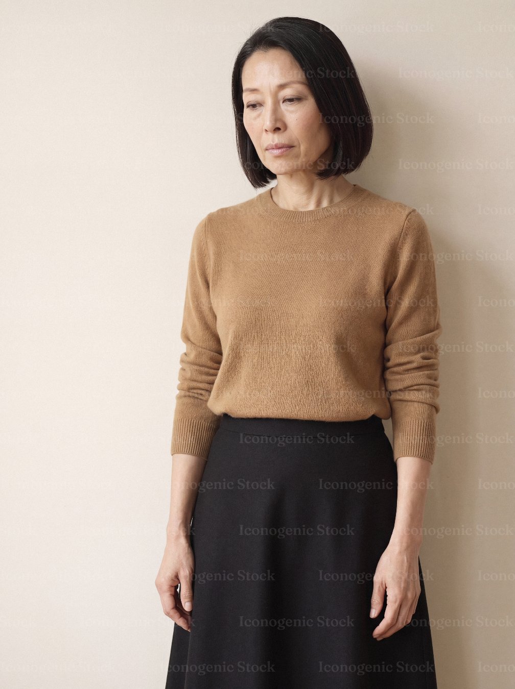 Middle-aged Asian woman in camel sweater standing thoughtfully against neutral wall
