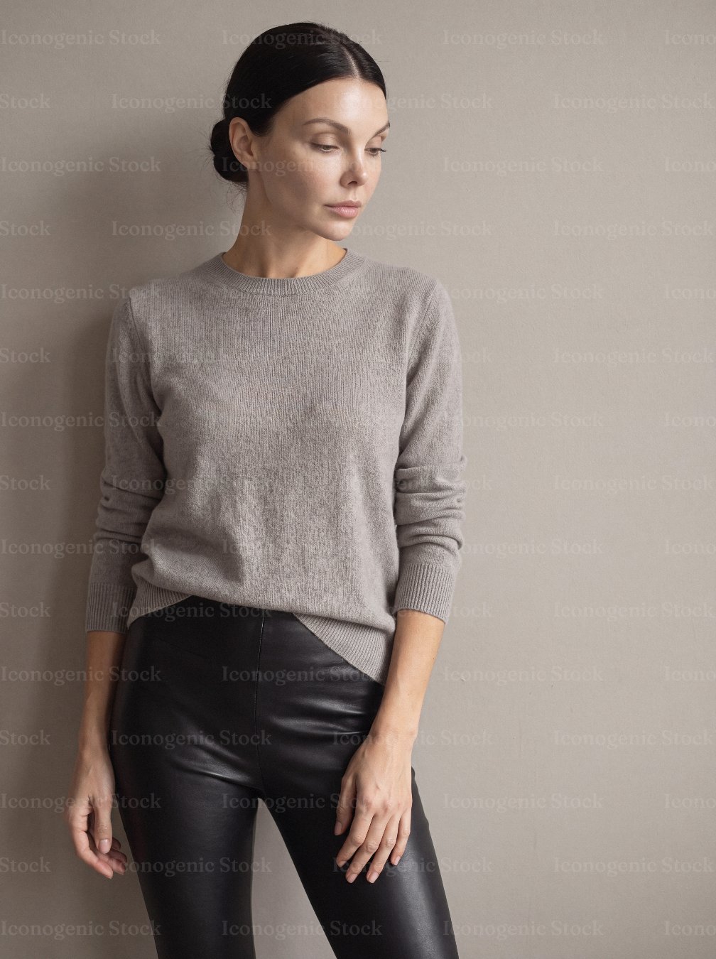 Woman in beige sweater and black leather pants posing against neutral wall
