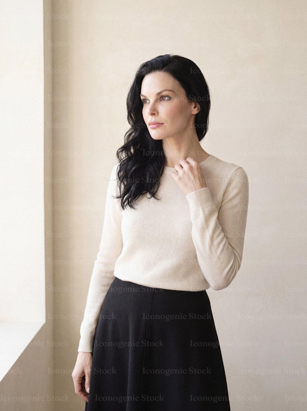 Elegant woman in cream sweater and black skirt standing by window, looking away