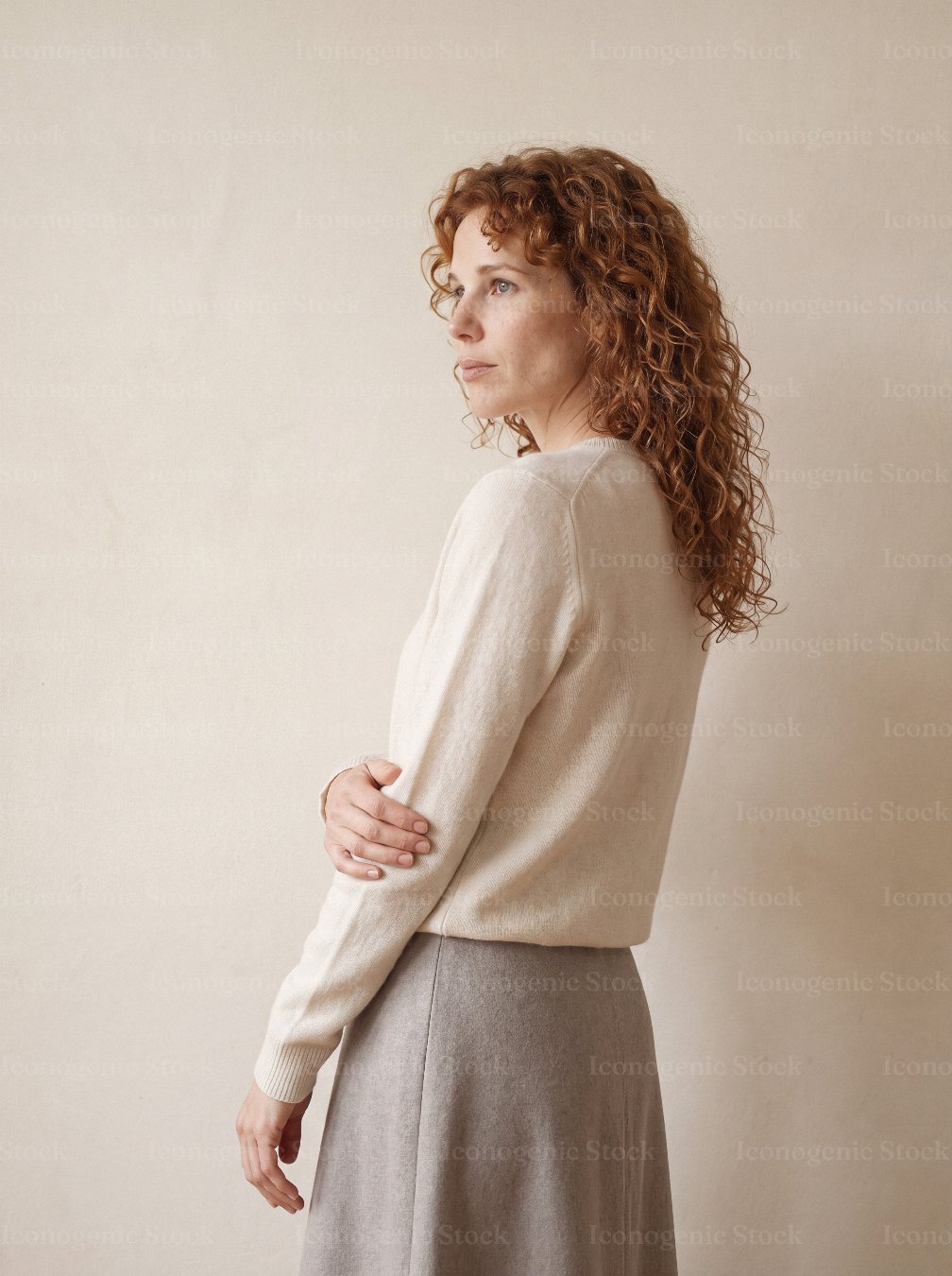 Redhead woman with curly hair in cream sweater looking over shoulder, minimalist studio portrait
