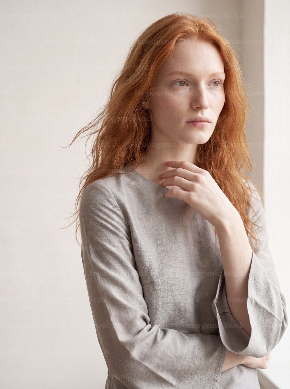Thoughtful redhead woman in linen blouse gazing out of window in soft light