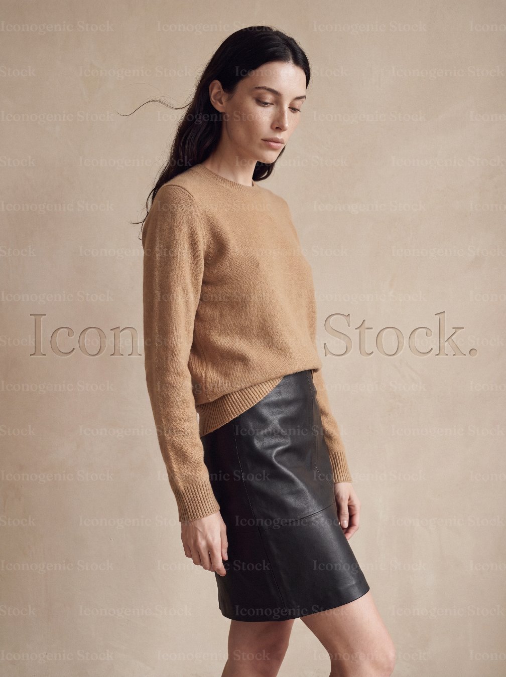 Woman in camel sweater and black leather skirt posing against neutral backdrop
