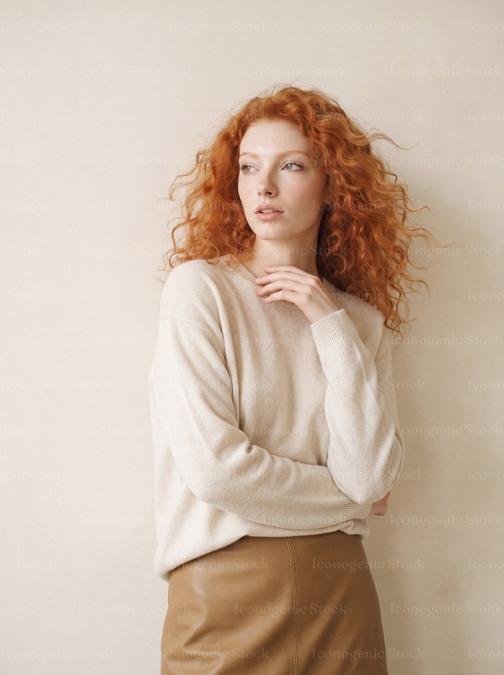 Redhead woman with curly hair in cream sweater posing thoughtfully against beige wall