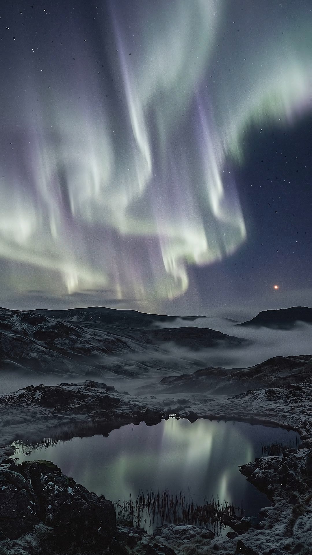 Vivid aurora borealis reflecting in still pond amid snowy mountain wilderness at night