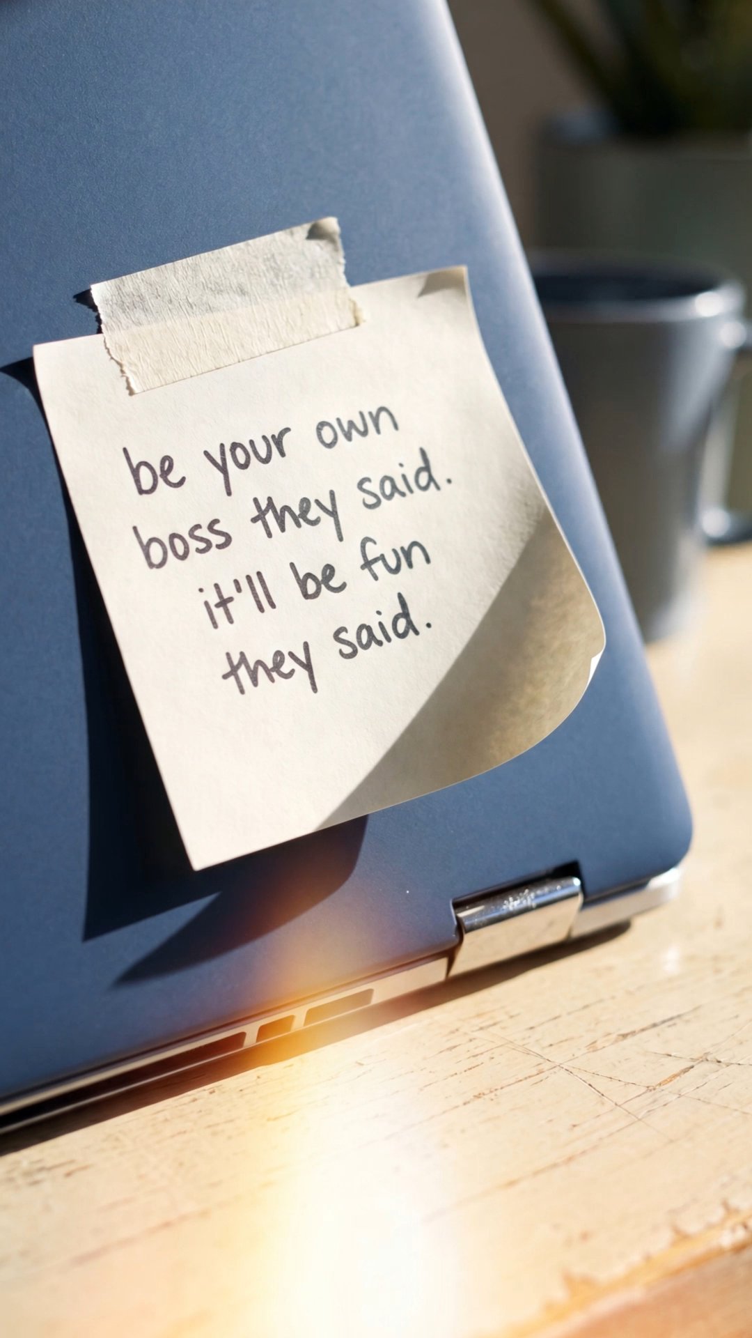 Sticky note reading 'be your own boss they said' taped to laptop on desk